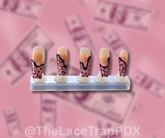 Five false nails glittry pink tips with pink and black cheetah print design on a clear stand against a pink background with dollar bill pattern.